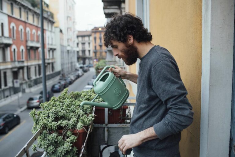 Ein junger Mann mit Bart gießt seine grünen Balkonpflanzen mit einer grünen Gießkanne auf einem Stadtbalkon