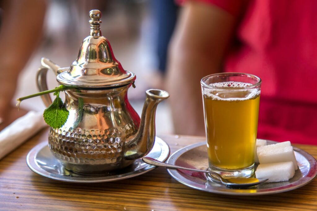 Eine traditionelle silberne Teekanne und ein Glas marokkanischer Minztee mit Schaumkrone und Zuckerstücken auf einem Holztisch